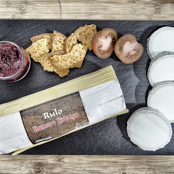 Rulo de cabra Romero Hidalgo con su etiqueta, rodajas de queso de cabra, tomate, mermelada y crujientes sobre pizarra